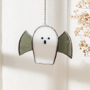 May include: A white stained glass ghost with gray wings, black eyes, and a small mouth. The ghost is hanging from a silver chain. The design is a Halloween decoration.