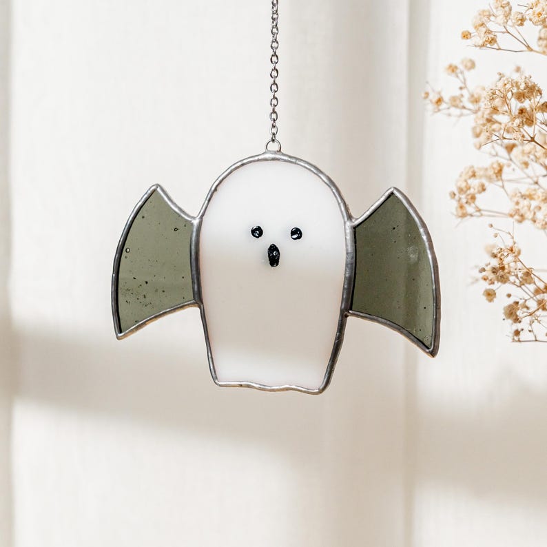 May include: A white stained glass ghost with gray wings, black eyes, and a small mouth. The ghost is hanging from a silver chain. The design is a Halloween decoration.