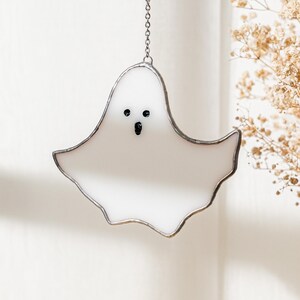 May include: A white stained glass ghost with a silver metal border and a chain for hanging. The ghost has two black dot eyes and a black oval mouth. The ghost is a Halloween decoration.