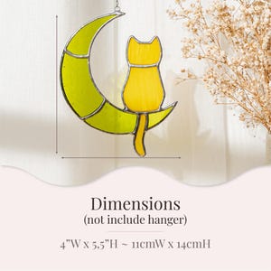 May include: A stained glass suncatcher featuring a yellow cat sitting on a crescent moon. The moon is green and the cat is yellow. The suncatcher is 4 inches wide and 5.5 inches high, or 11 centimeters wide and 14 centimeters high.