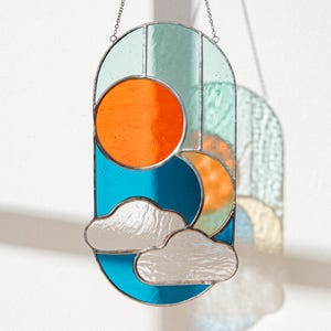 K&ouml;nnte beinhalten: Ein Buntglas-Sonnen- und Wolken-Design in den Farben T&uuml;rkis, Orange und Wei&szlig;. Das St&uuml;ck ist oval geformt und h&auml;ngt an einer silbernen Kette.