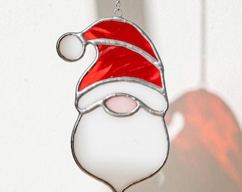 May include: A stained glass ornament shaped like a Santa Claus head, featuring a vibrant red hat with a white pom-pom, a white brim, a small pink nose, and a large white beard, all held together by silver metal solder lines and a chain.