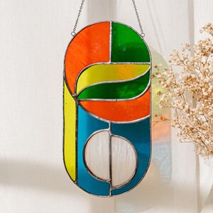 May include: A stained glass window hanging from a chain. The window is made of colourful glass pieces in shades of orange, yellow, green, and blue. The glass pieces are arranged in a geometric pattern.