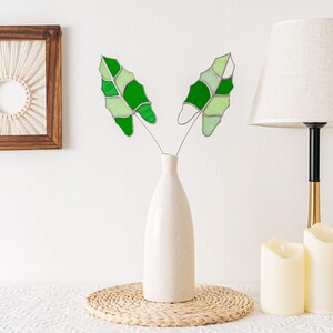 May include: A white ceramic vase holds two decorative glass leaves with shades of green. The leaves are on thin metal stems. The vase sits on a woven mat. A lamp and candles are in the background.