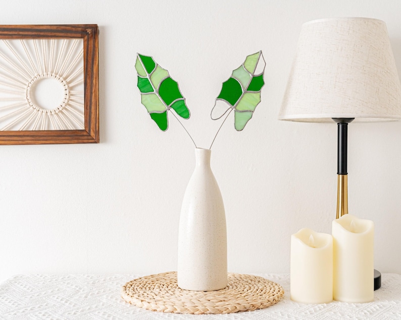 May include: A white ceramic vase holds two decorative glass leaves with shades of green. The leaves are on thin metal stems. The vase sits on a woven mat. A lamp and candles are in the background.