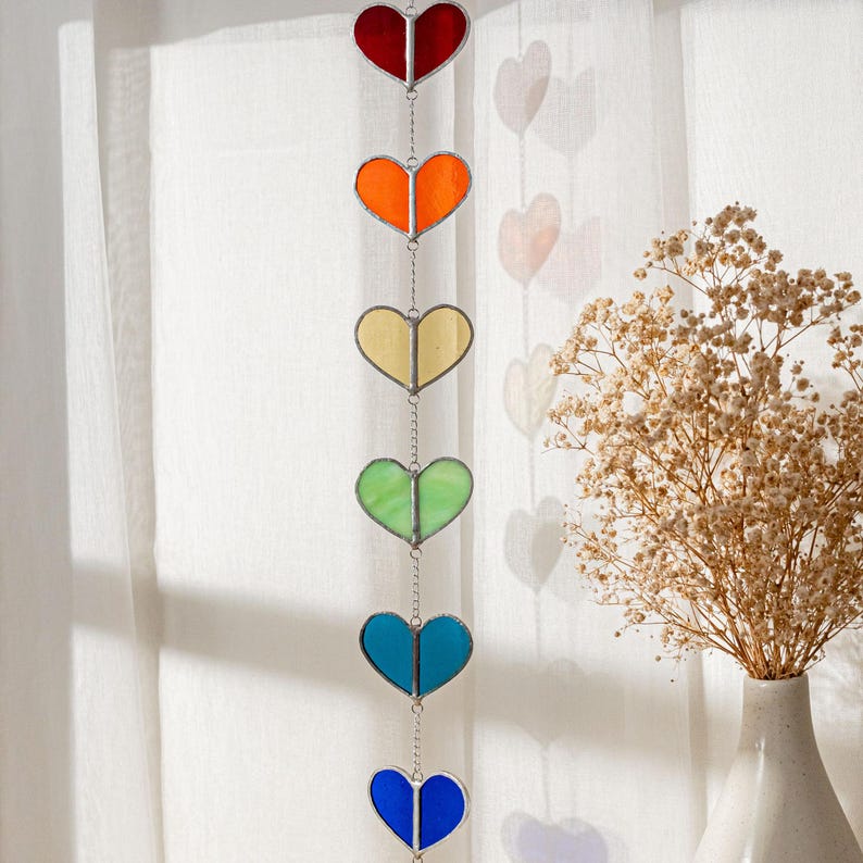 Op de afbeelding: Een hangende mobiel met zes gebrandschilderde glazen harten in regenboogkleuren: rood, oranje, geel, groen, blauw en indigo. De harten zijn verbonden door een zilveren ketting en hangen voor een wit gordijn.