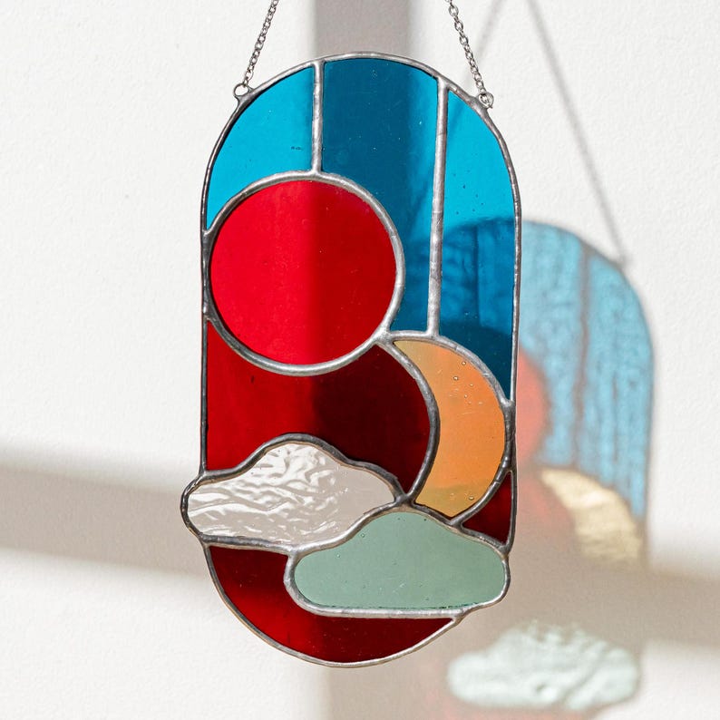 K&ouml;nnte beinhalten: Ein h&auml;ngendes Buntglasfenster mit Sonne und Mond in einem Farbschema aus Blau, Rot, Orange und Wei&szlig;. Sonne und Mond werden teilweise von wei&szlig;en Wolken verdeckt.