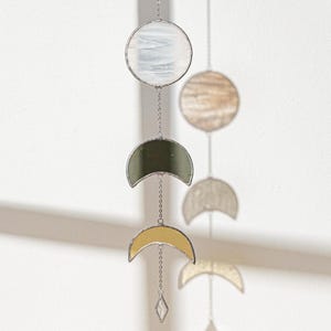 May include: A hanging stained glass moon phase mobile. The mobile features a series of crescent moons and circular shapes in various colors, including clear, olive green, and yellow. The pieces are connected by delicate chains and a diamond-shaped crystal.