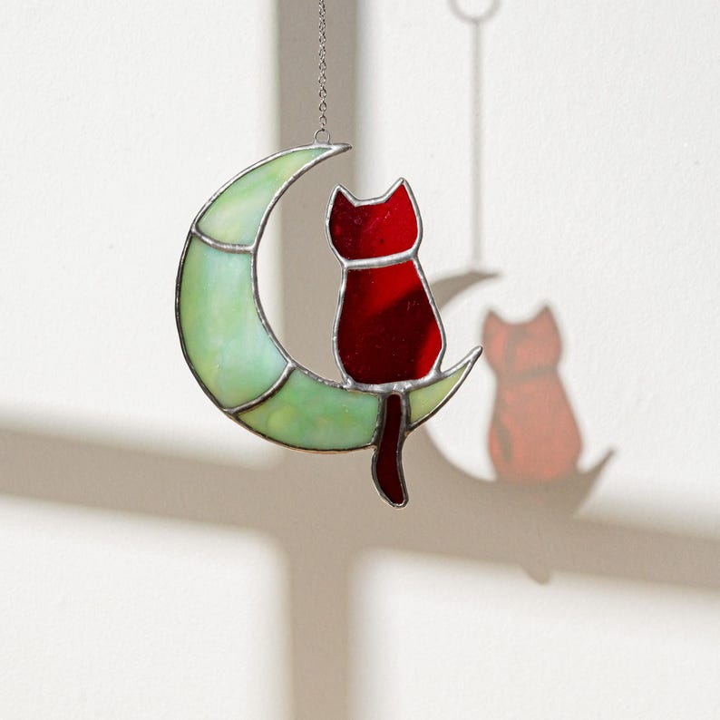 May include: A stained glass hanging ornament featuring a red cat sitting on a green crescent moon. The ornament is suspended from a silver chain.
