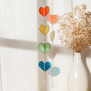 May include: A vertical stained glass suncatcher featuring four heart-shaped pieces in a gradient of orange, yellow, light green, and blue. Each heart is framed in silver metal and connected by a thin silver chain, ending with a small clear crystal.