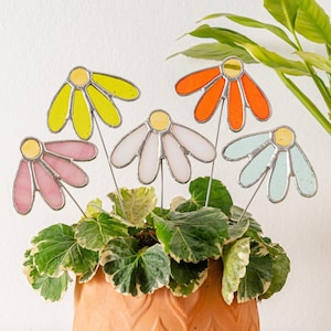 Op de afbeelding: Een verzameling kleurrijke glazen madeliefjes voor in de tuin. Elke bloem heeft een gele kern en bloemblaadjes in tinten geel, roze, wit, oranje en blauw. De stokken zijn in een pot met groen en wit gevlekt blad geplaatst.