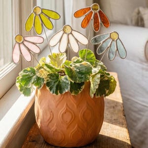May include: A terracotta ceramic pot with a patterned design contains a green and white leafy plant. Five colourful stained glass flower decorations are inserted into the plant. The flowers are yellow, orange, white, pink, and blue.
