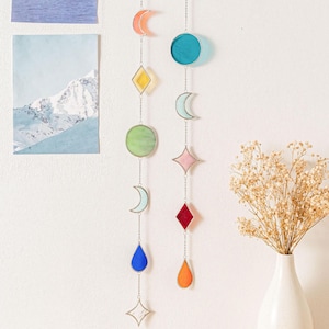 May include: Two hanging stained glass suncatchers with various shapes and colors, including moons, diamonds, circles, and teardrops. The suncatchers are suspended by silver chains and are displayed against a white wall. A vase of dried flowers is on the right.