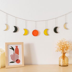 Puede incluir: Decoraci&oacute;n de pared con fases lunares amarillas, negras y naranjas suspendidas de una cadena plateada. Una impresi&oacute;n enmarcada y velas sobre una superficie de madera, contra una pared blanca.