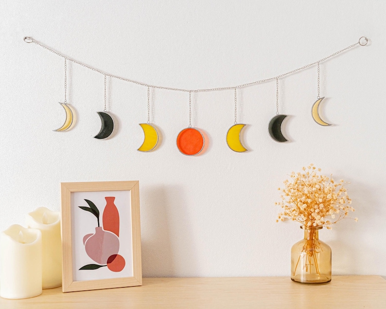Puede incluir: Decoraci&oacute;n de pared con fases lunares amarillas, negras y naranjas suspendidas de una cadena plateada. Una impresi&oacute;n enmarcada y velas sobre una superficie de madera, contra una pared blanca.