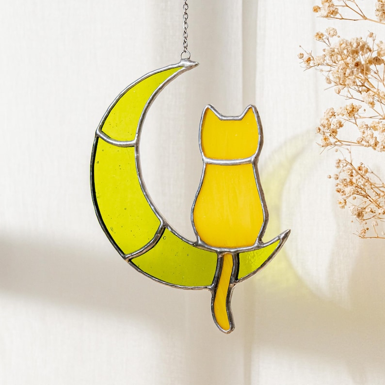 May include: A yellow stained glass cat sitting on a yellow crescent moon. The cat is facing away from the viewer and the moon is hanging from a silver chain.