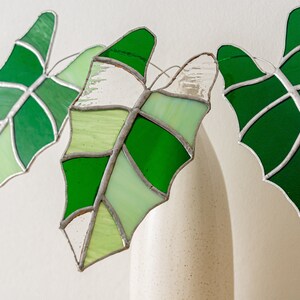 May include: Three stained glass leaf ornaments in shades of green and white. The leaves are outlined in silver and have a textured appearance. The leaves are hanging in front of a white vase.