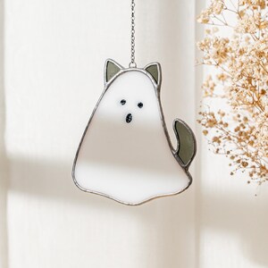 May include: A white and gray stained glass cat-shaped suncatcher hanging from a silver chain. The cat has black eyes and a tail. The cat's ears and tail are gray. The suncatcher is hanging in front of a white background.