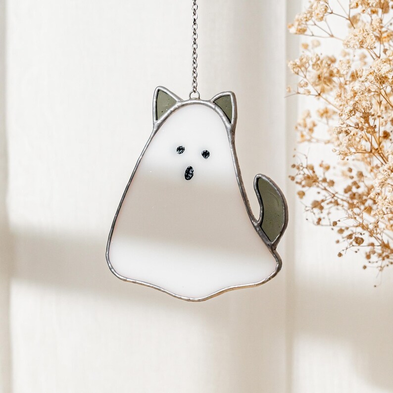 May include: A white and gray stained glass cat-shaped suncatcher hanging from a silver chain. The cat has black eyes and a tail. The cat's ears and tail are gray. The suncatcher is hanging in front of a white background.