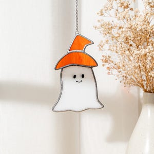 May include: A stained glass ghost decoration with an orange witch hat. The white ghost has a smiling face and is outlined in black. The Halloween-themed decoration hangs from a silver chain.
