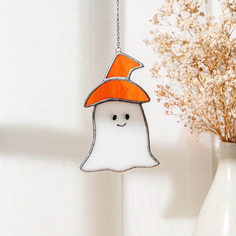 May include: A stained glass ghost decoration with an orange witch hat. The white ghost has a smiling face and is outlined in black. The Halloween-themed decoration hangs from a silver chain.