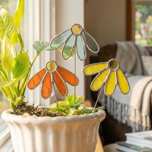 May include: Three colorful stained glass flower garden stakes in a white ceramic pot. The flowers are in shades of yellow, orange, and blue-green with silver-colored metal outlines. The pot sits in front of a window with a green plant.