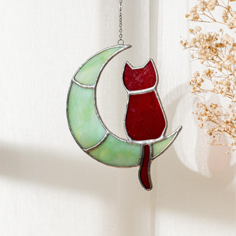 May include: A stained glass window hanging with a red cat sitting on a green crescent moon.
