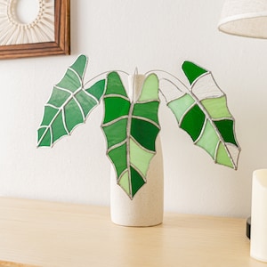 May include: A stained glass plant sculpture featuring three large green leaves in varying shades, set in a small, speckled white vase. The leaves have a detailed vein pattern and are displayed on a light-colored wooden surface.