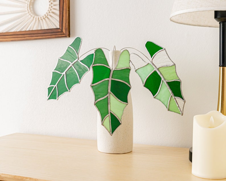 May include: A stained glass plant sculpture featuring three large green leaves in varying shades, set in a small, speckled white vase. The leaves have a detailed vein pattern and are displayed on a light-colored wooden surface.