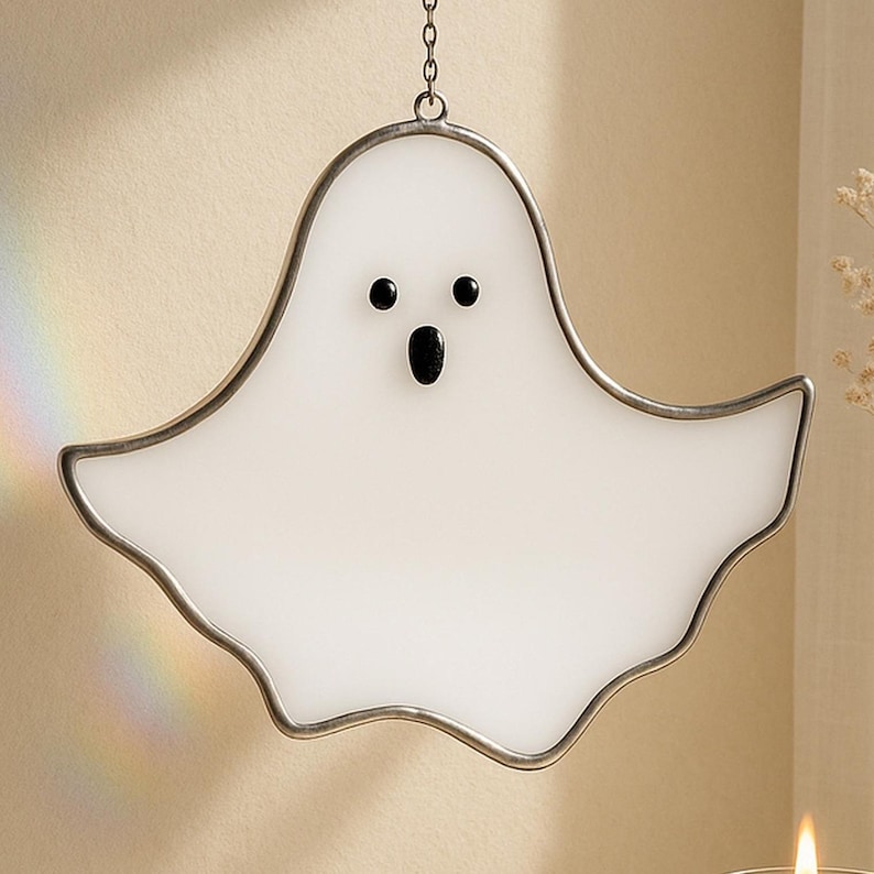 May include: A white ghost-shaped stained glass decoration with a silver metal frame and chain for hanging. The ghost has black eyes and a black open mouth. A decorative item for Halloween.