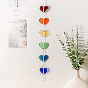 Op de afbeelding: Een hangend decoratief stuk met zeven hartvormige glas-in-lood panelen in regenboogkleuren: rood, oranje, geel, groen, blauw en indigo. De harten zijn verbonden door een zilveren ketting en hangen verticaal.