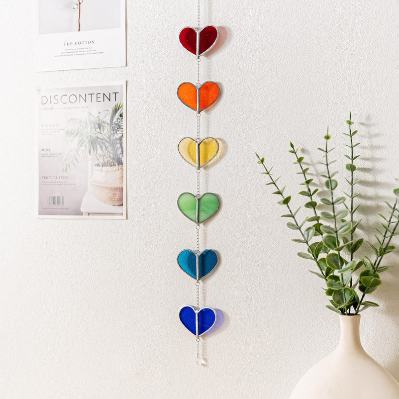 Op de afbeelding: Een hangend decoratief stuk met zeven hartvormige glas-in-lood panelen in regenboogkleuren: rood, oranje, geel, groen, blauw en indigo. De harten zijn verbonden door een zilveren ketting en hangen verticaal.