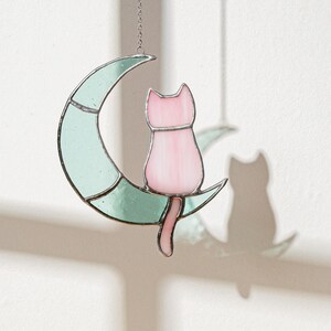 May include: A stained glass cat sitting on a crescent moon. The cat is pink and the moon is light green.