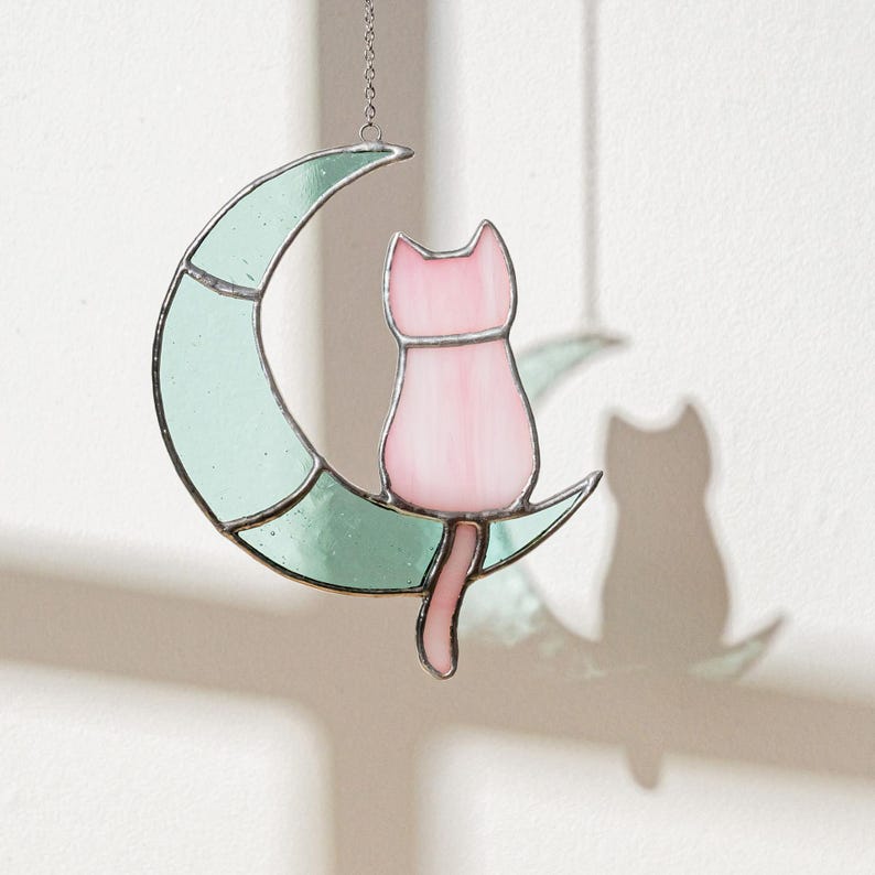 May include: A stained glass cat sitting on a crescent moon. The cat is pink and the moon is light green.