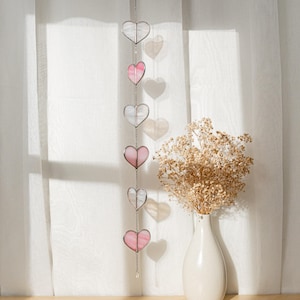 Op de afbeelding: Een decoratief hangend hartornament met zes glazen harten in afwisselende roze en heldere kleuren. De harten zijn verbonden door een zilveren ketting en hangen voor een wit gordijn. Een vaas met droogbloemen staat op de achtergrond.