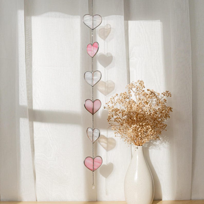 Op de afbeelding: Een decoratief hangend hartornament met zes glazen harten in afwisselende roze en heldere kleuren. De harten zijn verbonden door een zilveren ketting en hangen voor een wit gordijn. Een vaas met droogbloemen staat op de achtergrond.