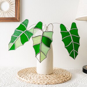 May include: Stained glass leaves in shades of green, arranged in a cream-colored vase. The leaves have a detailed vein pattern and are displayed on a white surface with a woven placemat. A decorative piece for home decor.