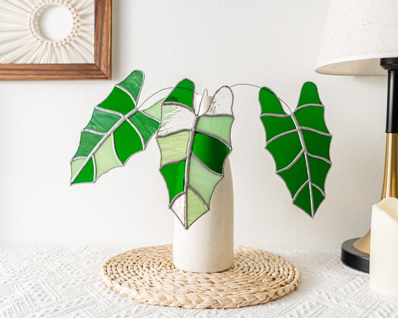 May include: Stained glass leaves in shades of green, arranged in a cream-colored vase. The leaves have a detailed vein pattern and are displayed on a white surface with a woven placemat. A decorative piece for home decor.