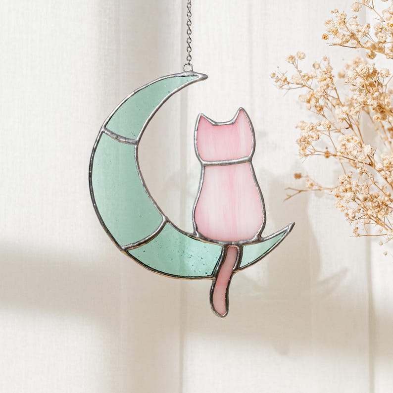 May include: A stained glass window hanging with a pink cat sitting on a crescent moon. The moon is a light green color.
