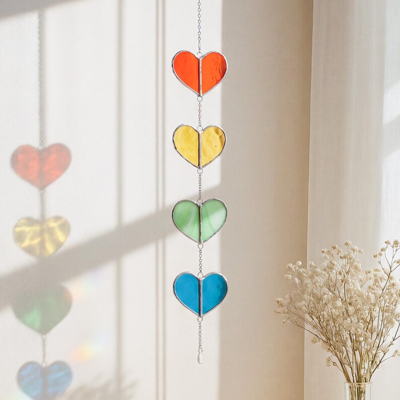 Op de afbeelding: Een hangende gebrandschilderde glazen hartdecoratie met vier harten in rood, geel, groen en blauw. Elk hart is omrand met zilver en verbonden door een ketting. Een kleine, heldere kraal hangt aan de onderkant.