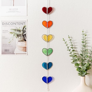 May include: A decorative hanging ornament featuring six heart-shaped stained glass panels in a rainbow of colours: red, orange, yellow, green, blue, and indigo. The hearts are linked by a silver chain and hang against a white wall.