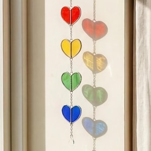 Op de afbeelding: Een kleurrijke hartvormige zonnevanger met rode, gele, groene en blauwe glazen panelen. De harten zijn verbonden door een fijne ketting en een kleine kristal aan de onderkant. Ontworpen om in een raam te hangen.
