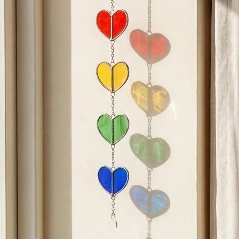 Op de afbeelding: Een kleurrijke hartvormige zonnevanger met rode, gele, groene en blauwe glazen panelen. De harten zijn verbonden door een fijne ketting en een kleine kristal aan de onderkant. Ontworpen om in een raam te hangen.