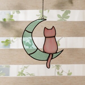 May include: A stained glass cat sitting on a crescent moon. The cat is pink and the moon is green.