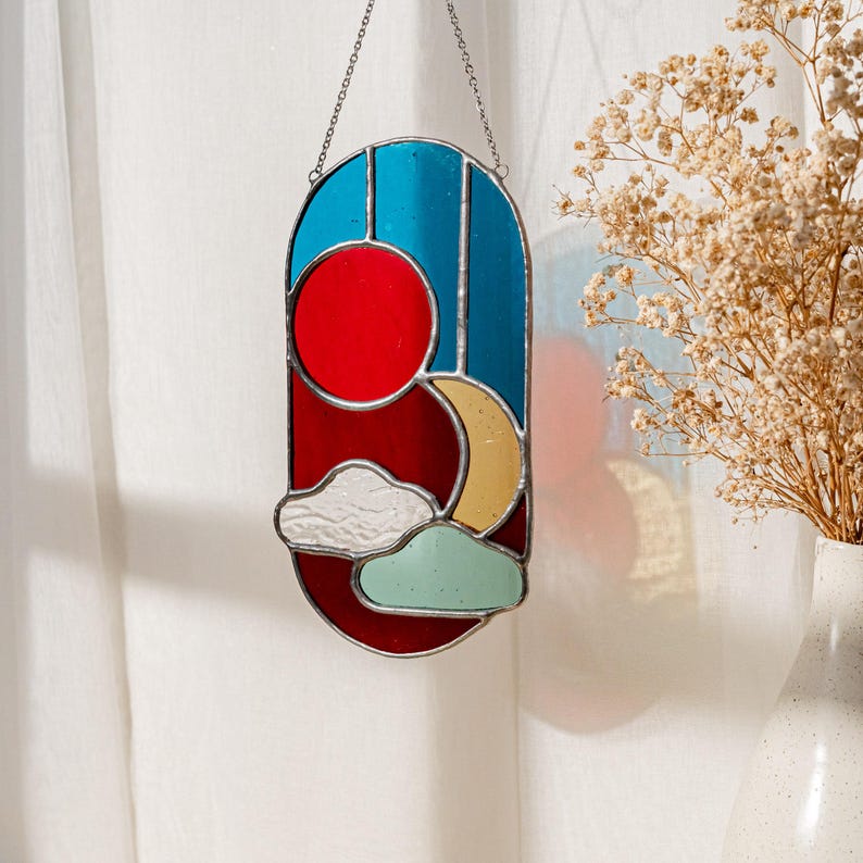 K&ouml;nnte beinhalten: Ein Buntglas-Sonnenf&auml;nger mit einer roten Sonne, einem gelben Halbmond und blauen und wei&szlig;en Wolken. Der Sonnenf&auml;nger h&auml;ngt an einer silbernen Kette.
