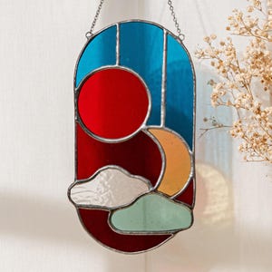K&ouml;nnte beinhalten: Ein Buntglasfenster mit einem Sonnen- und Mondmotiv in den Farben Rot, Orange, Blau und Gr&uuml;n. Sonne und Mond sind von Wolken und einem roten Hintergrund umgeben.
