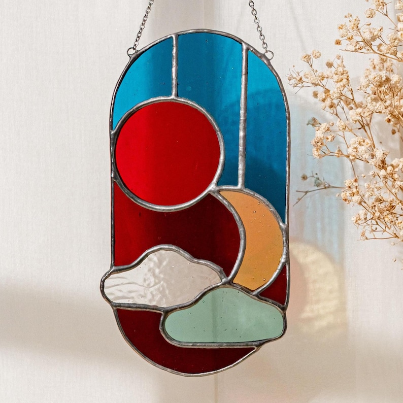 K&ouml;nnte beinhalten: Ein Buntglasfenster mit einem Sonnen- und Mondmotiv in den Farben Rot, Orange, Blau und Gr&uuml;n. Sonne und Mond sind von Wolken und einem roten Hintergrund umgeben.
