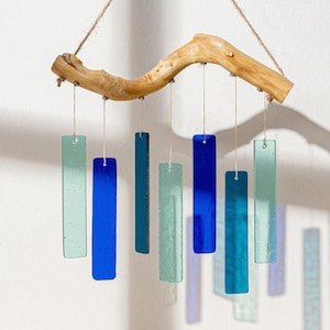 Puede incluir: Un carillón de viento artesanal con piezas rectangulares de vidrio en tonos azules y verdes suspendidas de una rama de madera natural. Las campanillas cuelgan de finos cordones, creando un acento decorativo y relajante.