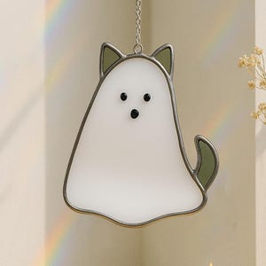 May include: A white ghost-shaped stained glass suncatcher with black dot eyes and a green tail and ears. The suncatcher is outlined in a silver metal frame and hangs from a silver chain. A rainbow is visible in the background.