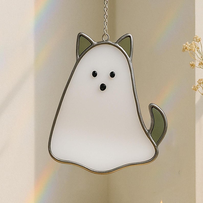 May include: A white ghost-shaped stained glass suncatcher with black dot eyes and a green tail and ears. The suncatcher is outlined in a silver metal frame and hangs from a silver chain. A rainbow is visible in the background.